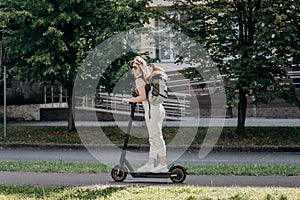 Happy smiling woman traveler is riding her electro scooter in city parkland with dog Welsh Corgi Pembroke in a special backpack