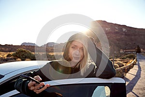 Happy smiling woman using smartphone with online map, enjoying a car road trip along the mountainous landscape during sunrise