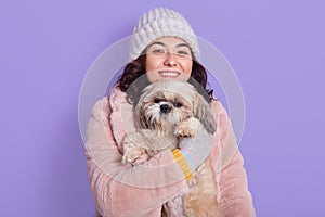 Happy smiling woman wearing faux fur coat and warm white cap, hugging Pekingese dog, posing isolated over lilac background,