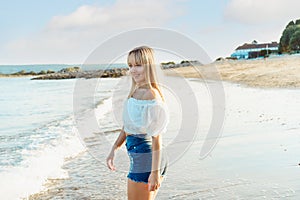 Happy smiling woman in white top and jeans shorts enjoying the moment during a walk on the seaside beach. Summer