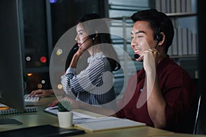 Happy smiling young Asian man with headphones working at call center service desk consultant with her teammates at night, taking