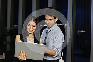 Happy smiling young Asian man and woman teammate officer have overwork project overnight in office, use laptop computer for