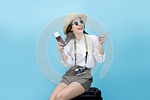 Happy smiling young Asian tourist woman sitting on luggage holding passport and hand pointing to copy space going to travel on
