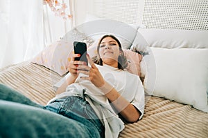 Happy smiling young attractive caucasian woman in eyeglasses using smartphone, lying on a bed
