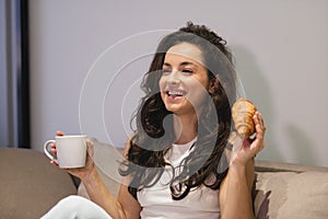 Happy smiling young attractive female resting at home holding in hands cup of tea and croissant. Close up concept