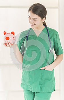 Happy smiling young beautiful female doctor with stethoscope holding pink piggy bank