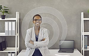 Happy smiling young black businesswoman standing with arms folded by office desk