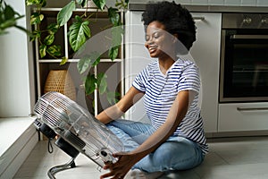 Happy smiling young black woman enjoying fresh air while cooling by electric fan at home