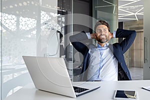 Happy and smiling young businessman man sitting in the office at the desk and resting and satisfied with the work with