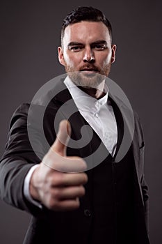 Happy smiling young businessman with raised thumb gesture, photo on dark background