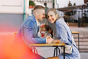 Happy smiling young couple meeting on a date enjoys life sitting at a table, holding hands, in a street cafe on a summer day