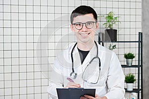 Happy smiling young doctor writing on clipboard in a modern hospital