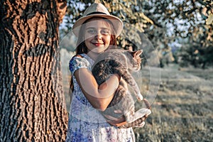 Happy smiling young girl in white hat and dress with flowers print holding a white and grey cat on her hands, looking at