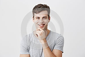 Happy smiling young man demonstrates positive emotions or feelings, has trendy hairdo, dressed casually, keeps hand on