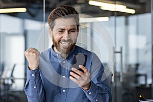 Happy and smiling young man sitting in office modern center and looking at phone, enjoying success and showing victory