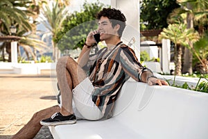 Happy smiling young man sitting outdoors with joyful expression on face during talking by mobile phone