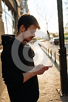 Happy smiling young man using modern smart phone while standing at crosswalk at the sunset in city center