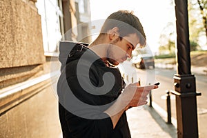 Happy smiling young man using modern smart phone while standing at crosswalk at the sunset in city center