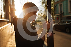 Happy smiling young man using modern smart phone while standing at crosswalk at the sunset in city center