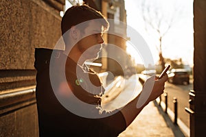 Happy smiling young man using modern smart phone while standing at crosswalk at the sunset in city center