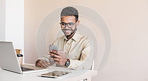 Happy smiling young man using smart phone and laptop at office. Handsome businessman working on computer and  texting phone