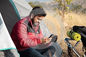 Happy smiling young Traveler busy using mobile phone from camping tent - Concept of Digital nomadic lifestyle, traveling and