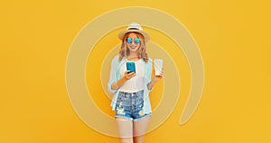 Happy smiling young woman 20s with smartphone and cup of coffee wearing summer straw hat, shorts on orange background