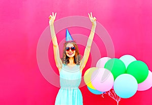 Happy smiling young woman in a birthday cap having fun on an air colorful balloons pink background