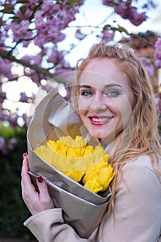Happy smiling young woman with blue eyes holding bouquets of yellow tulips, blossoming tree on background