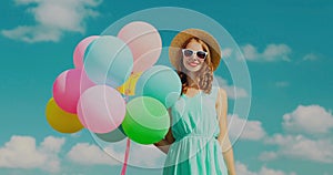 Happy smiling young woman with bunch of colorful balloons wearing a summer straw hat on the field on blue sky background