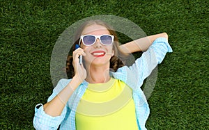 Happy smiling young woman calling on mobile phone while lying on grass in summer park