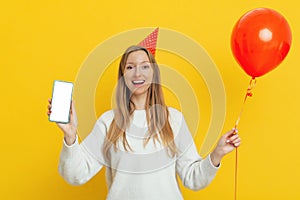 Happy smiling young woman in celebratory cap holding cell phone with empty screen for ad, wearing white casual style sweater,