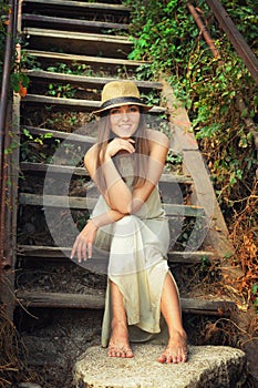 Happy smiling young woman dressed in hat and white long dress sitting barefoot on a vintage wooden stairs.