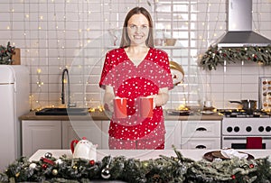 Happy smiling young woman holding or carrying two cups or mugs of tea at kitchen at christmas time