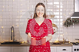 Happy smiling young woman holding or carrying two cups or mugs of tea at kitchen at christmas time