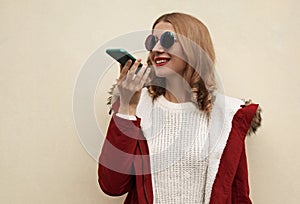 Happy smiling young woman holding smartphone using voice command recorder, assistant or takes calling wearing red jacket