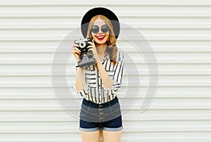 Happy smiling young woman holding vintage film camera in black round hat, shorts, white striped shirt on white wall