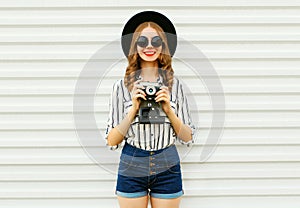 Happy smiling young woman holding vintage film camera in black round hat, shorts, white striped shirt on white wall