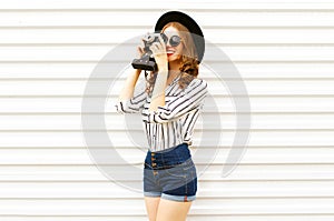 Happy smiling young woman holding vintage film camera in black round hat, shorts, white striped shirt on white wall