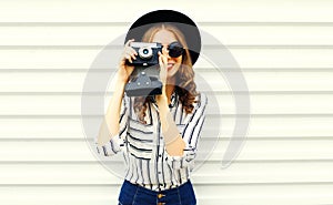 Happy smiling young woman holding vintage film camera in black round hat, shorts, white striped shirt on white wall