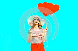 Happy smiling young woman with lollipop, red heart shaped air balloons in summer straw hat and shorts on colorful blue