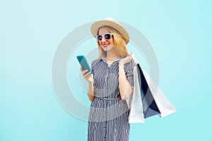 Happy smiling young woman with phone, holding colorful shopping bags in summer round straw hat, striped dress on blue