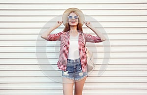 Happy smiling young woman posing in summer round straw hat, checkered shirt, shorts on white wall