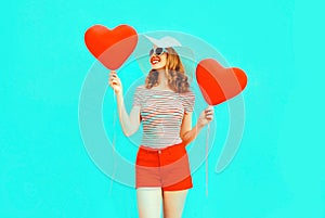 Happy smiling young woman with red heart shaped air balloons in summer straw hat and shorts on colorful