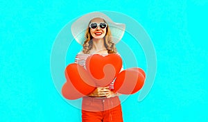 Happy smiling young woman with red heart shaped air balloons in summer straw hat and shorts on colorful blue