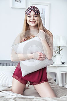 Happy smiling young woman sitting in her bed at home with pillow in her hands, wearing nighty and sleeping eye mask.