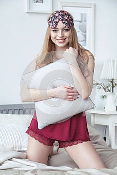 Happy smiling young woman sitting in her bed at home, wearing nighty and sleeping eye mask.