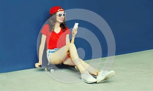 Happy smiling young woman with smartphone sitting on skateboard wearing red baseball cap, shorts on city street on blue wall