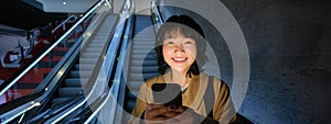 Happy smiling young woman, standing on escalator, going down, holding smartphone in both hands, chatting on mobile phone