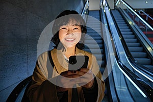 Happy smiling young woman, standing on escalator, going down, holding smartphone in both hands, chatting on mobile phone